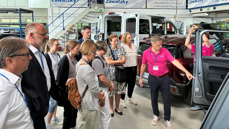 In einer Fahrzeughalle steht ein Mann vor einem Auto und zeigt auf die geöffnete Wagentür; eine Gruppe verschiedener Menschen hört aufmerksam zu.