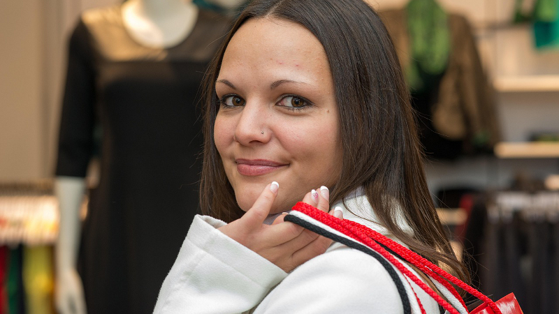 Eine junge Frau mit dunklen Haaren steht vor einem Bekleidungsgeschäft. Sie hat mehrere Einkaufstaschen locker über die Schulter gelegt und lächelt in die Kamera.